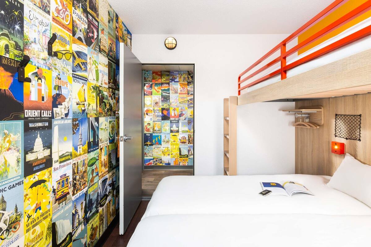 Une chambre avec un lit superposé, un mur décoré d’affiches de voyage, une porte ornée d’un globe terrestre et une petite table sur laquelle se trouvent un livre et un téléphone.