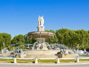 Une fontaine classique ornée de statues, entourée d’une clôture en treillis métallique, avec des arbres en arrière-plan.