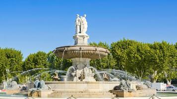 A classical fountain with statues, surrounded by a chain-link fence, and trees in the background.