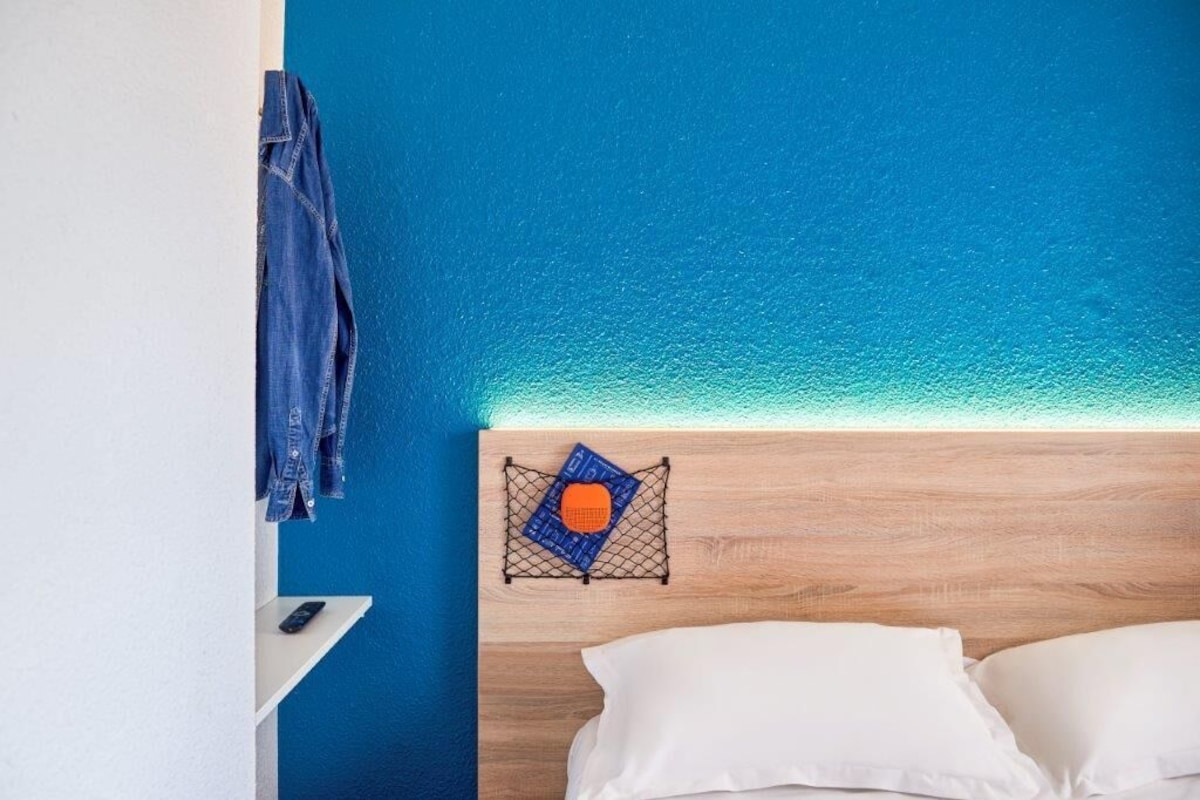 Een kamer met een houten hoofdbord, een bed met witte kussens, een blauwe wand en een hangend denim jasje.