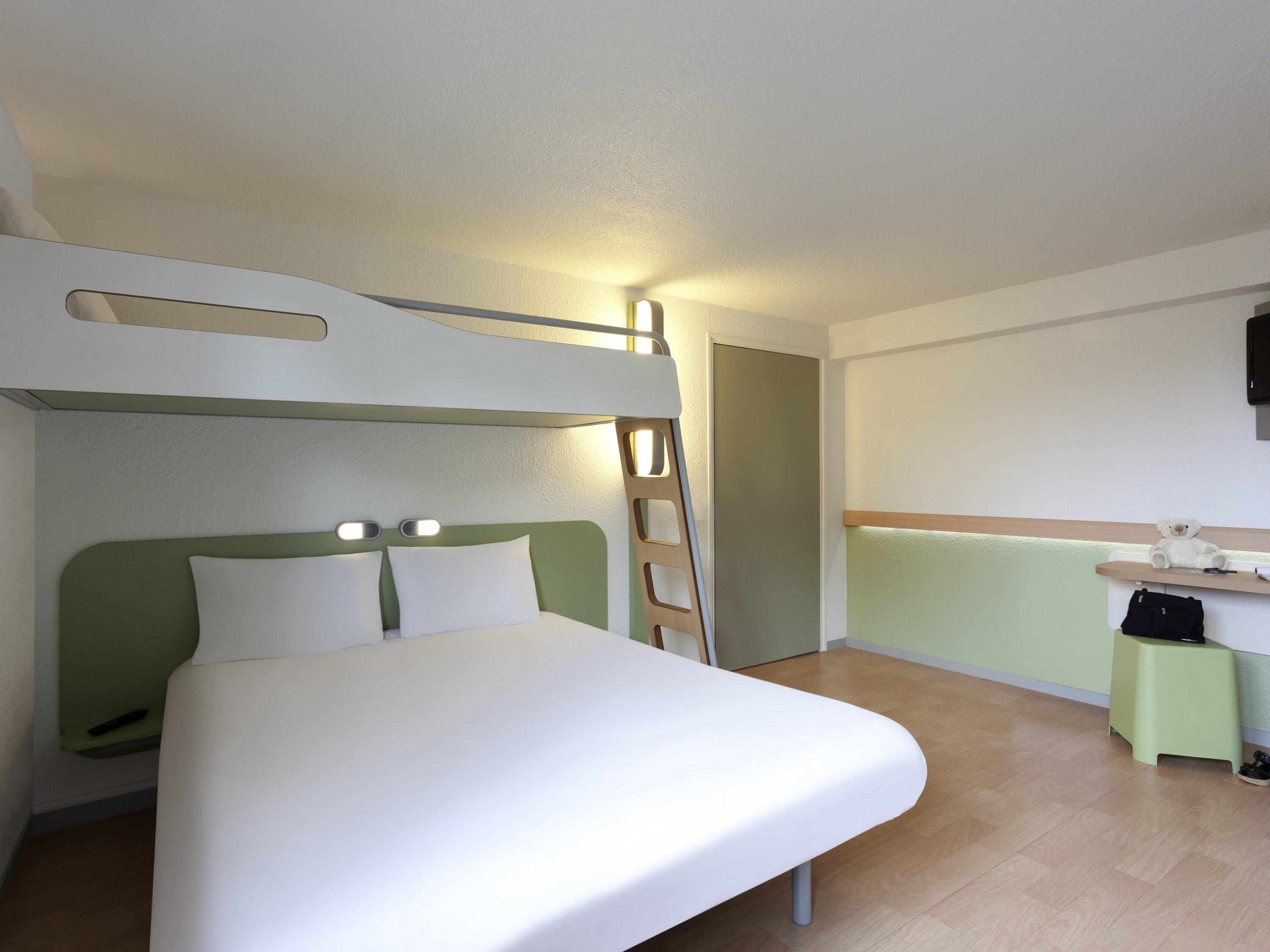 A room with a bunk bed, a desk with a chair, a small stool, and a television mounted on the wall.