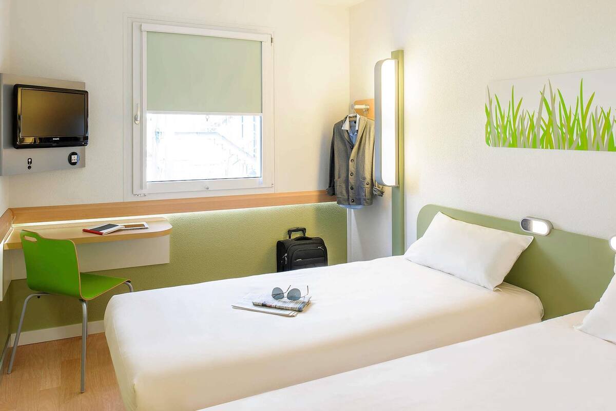 A hotel room with two beds, a desk, a chair, a TV, and a window with a view of buildings.