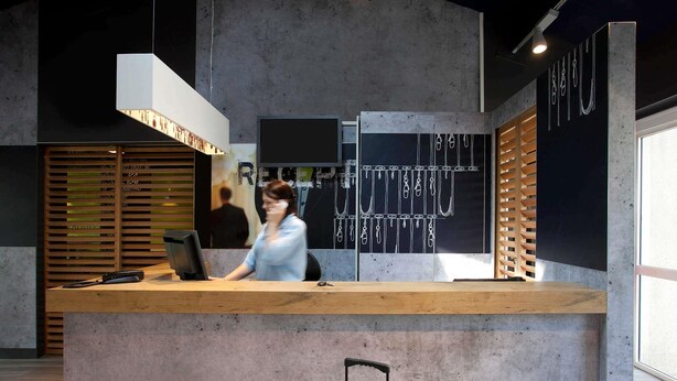 A reception desk with a wooden counter, a hanging light fixture, and a wall with jewelry displays. - ibis budget Tarbes (Tarbes)