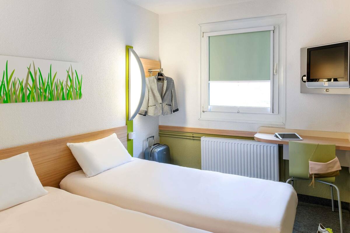 A hotel room with two beds, a desk with a computer, a TV, and a window.