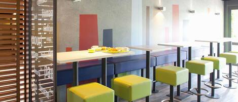 Interior de una cafetería moderna con una barra larga, taburetes coloridos y una pared con diseños abstractos.