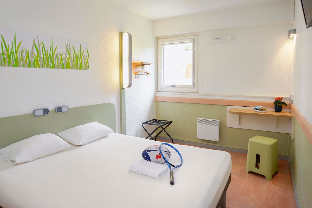 Ein ordentlich bezogenes Bett mit weißer Decke und Kissen, ein Nachttisch mit einem Buch und einer Topfpflanze, ein Fenster mit Blick nach draußen sowie ein Wandregal mit einem aufgehängten Kleidungsstück.