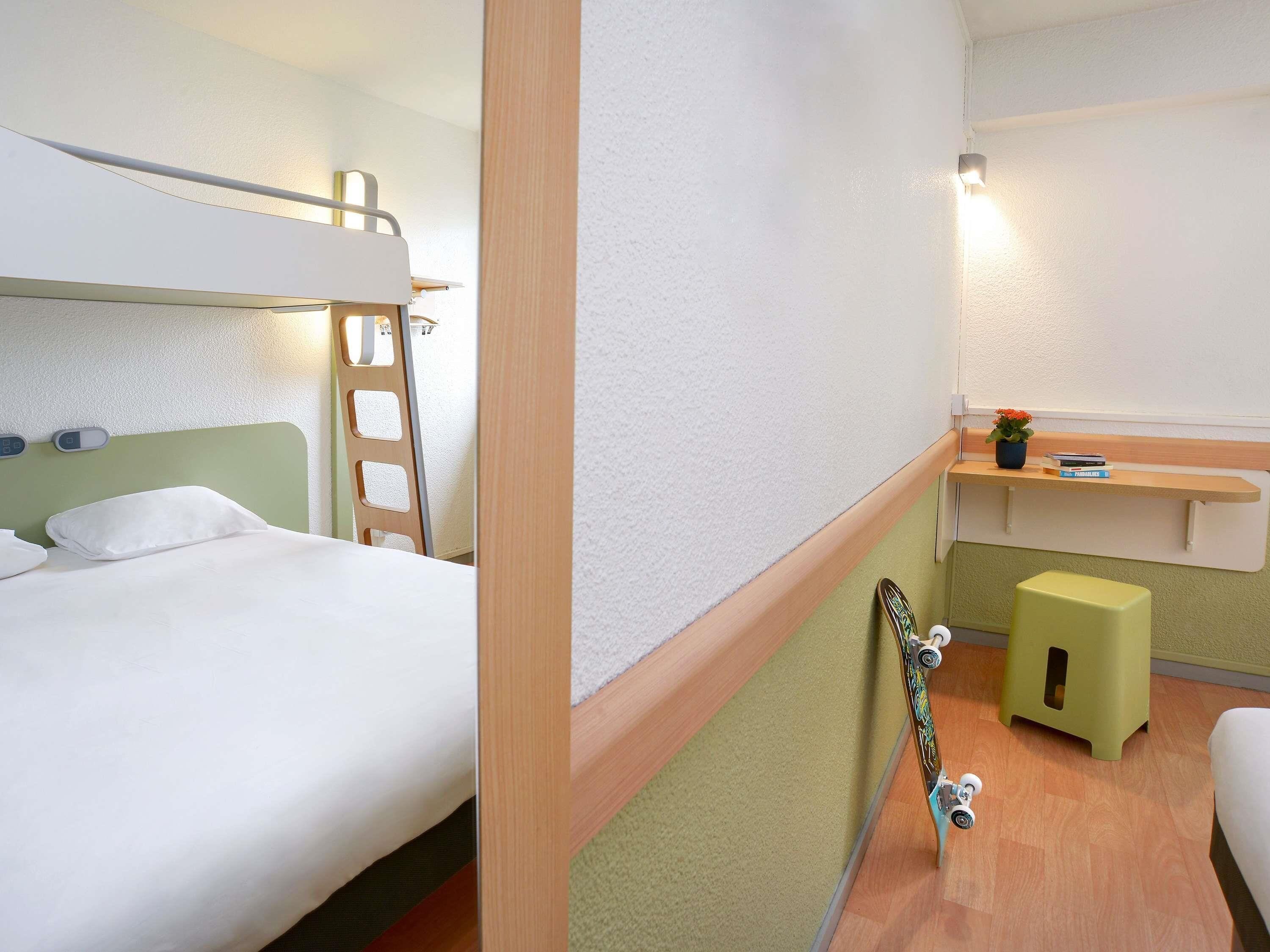 A compact hotel room with bunk beds, a small desk with a stool, a skateboard, and a potted plant.