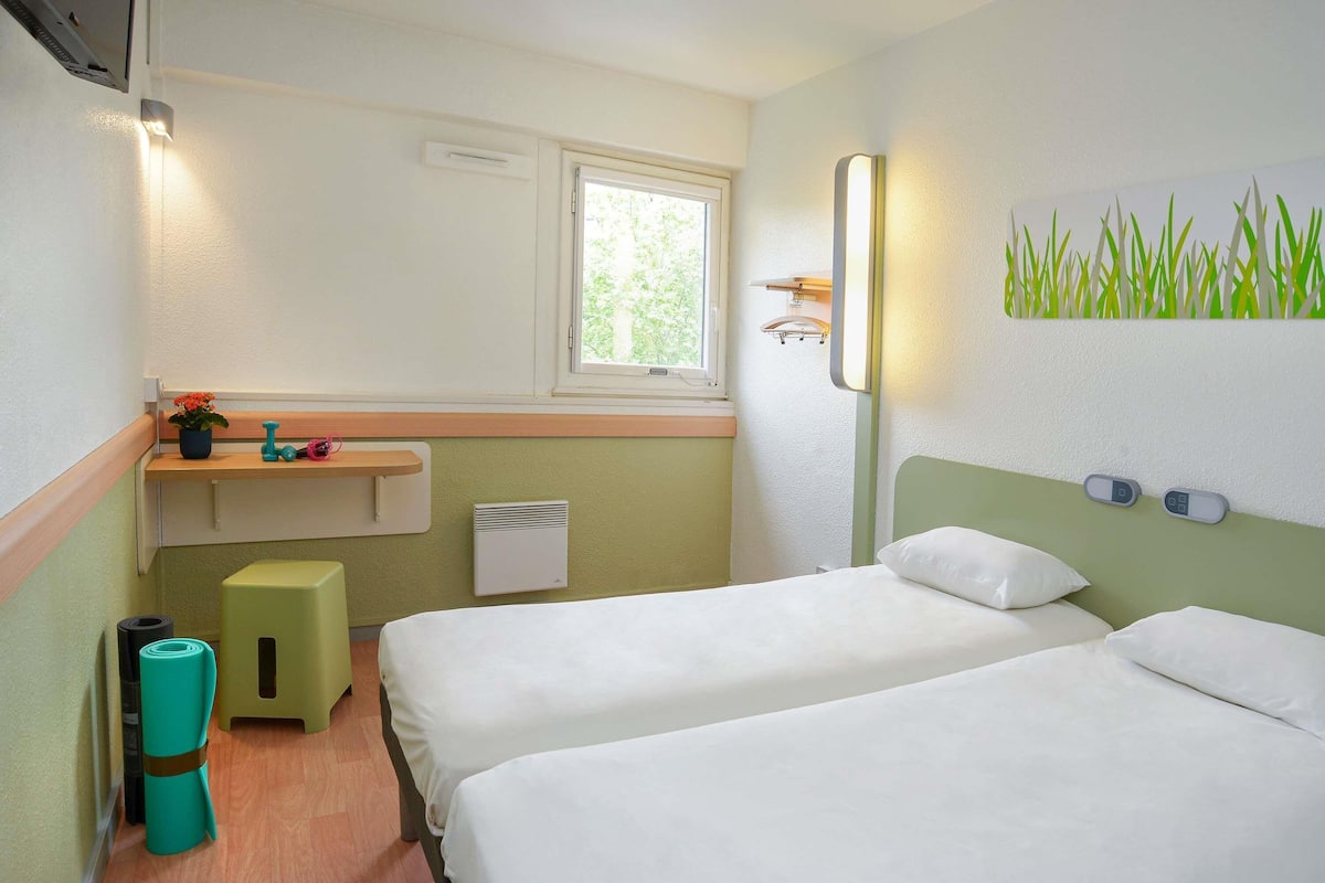 Ein Hotelzimmer mit zwei Betten, einem Fenster, einem kleinen Waschbecken, einem Heizkörper und einer Wandleuchte.