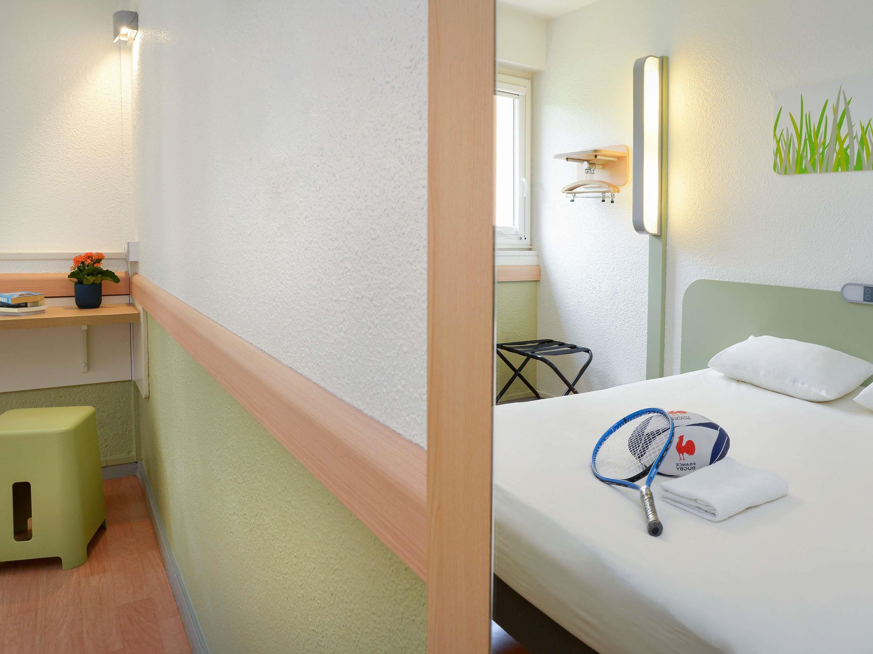 A hotel room with a bed, a small desk with a plant, a wall-mounted shelf, and a window.