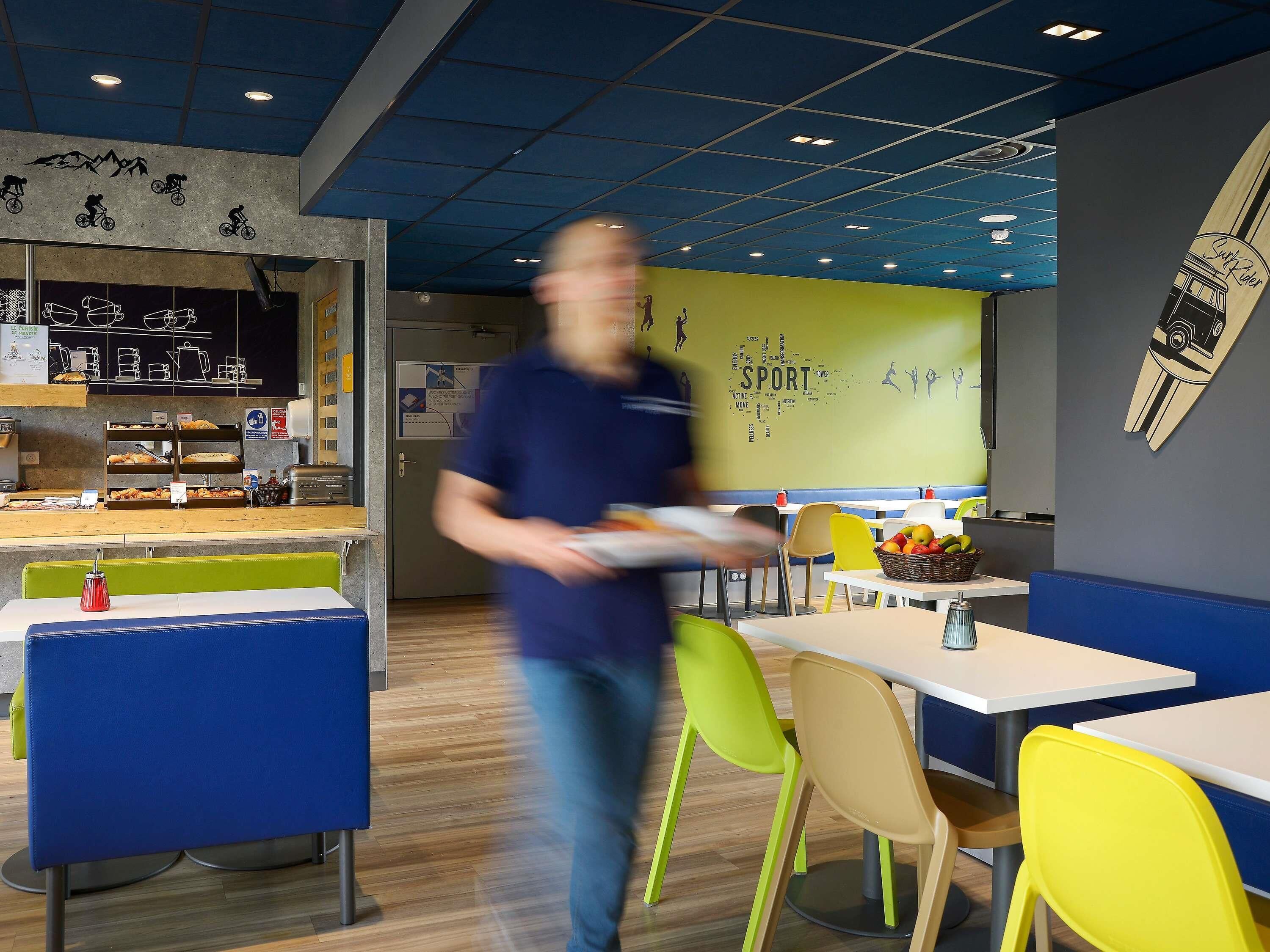 Un café moderno con una pared decorada con motivos deportivos, mesas de madera y sillas coloridas.