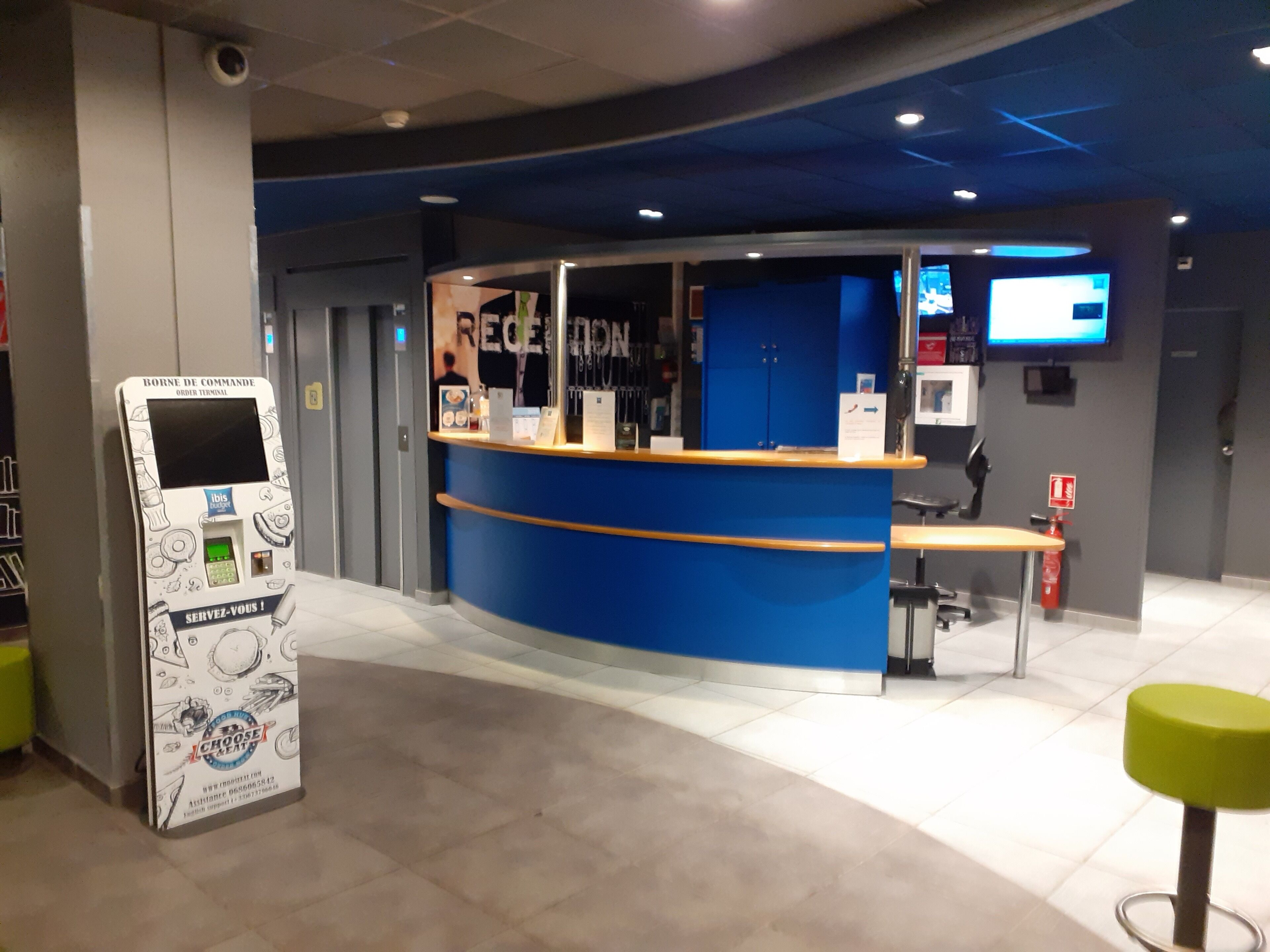 A reception area with a blue counter, a digital kiosk, and a seating area with a green stool.