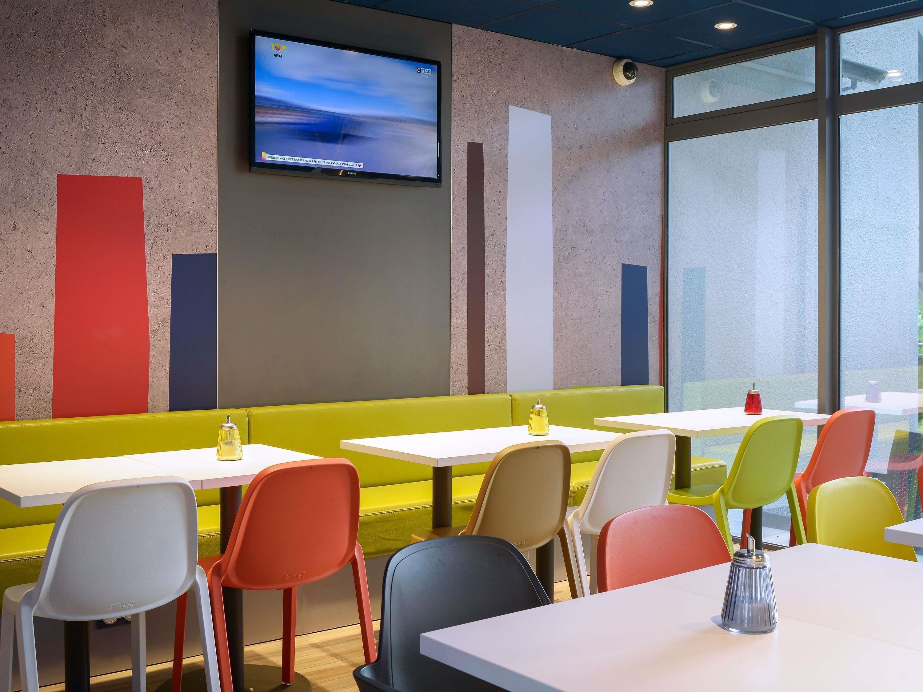 L’intérieur d’un restaurant moderne est agrémenté d’un téléviseur à écran plat fixé au mur, de panneaux verticaux aux couleurs vives et d’un assortiment de chaises blanches et colorées disposées autour des tables.