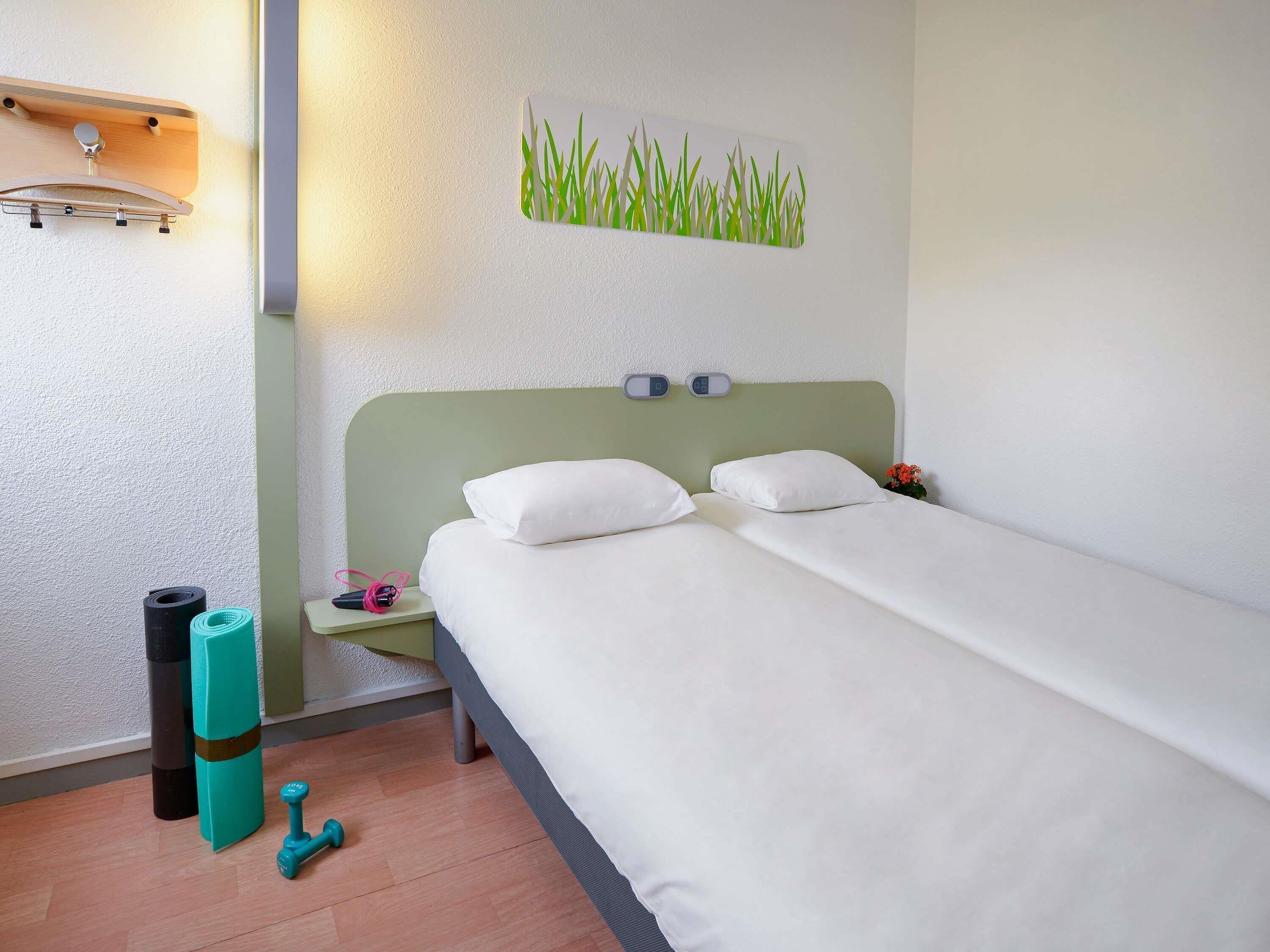 A hotel room with a bed, a wall-mounted shelf, a small table with a lamp, and a picture of grass on the wall.