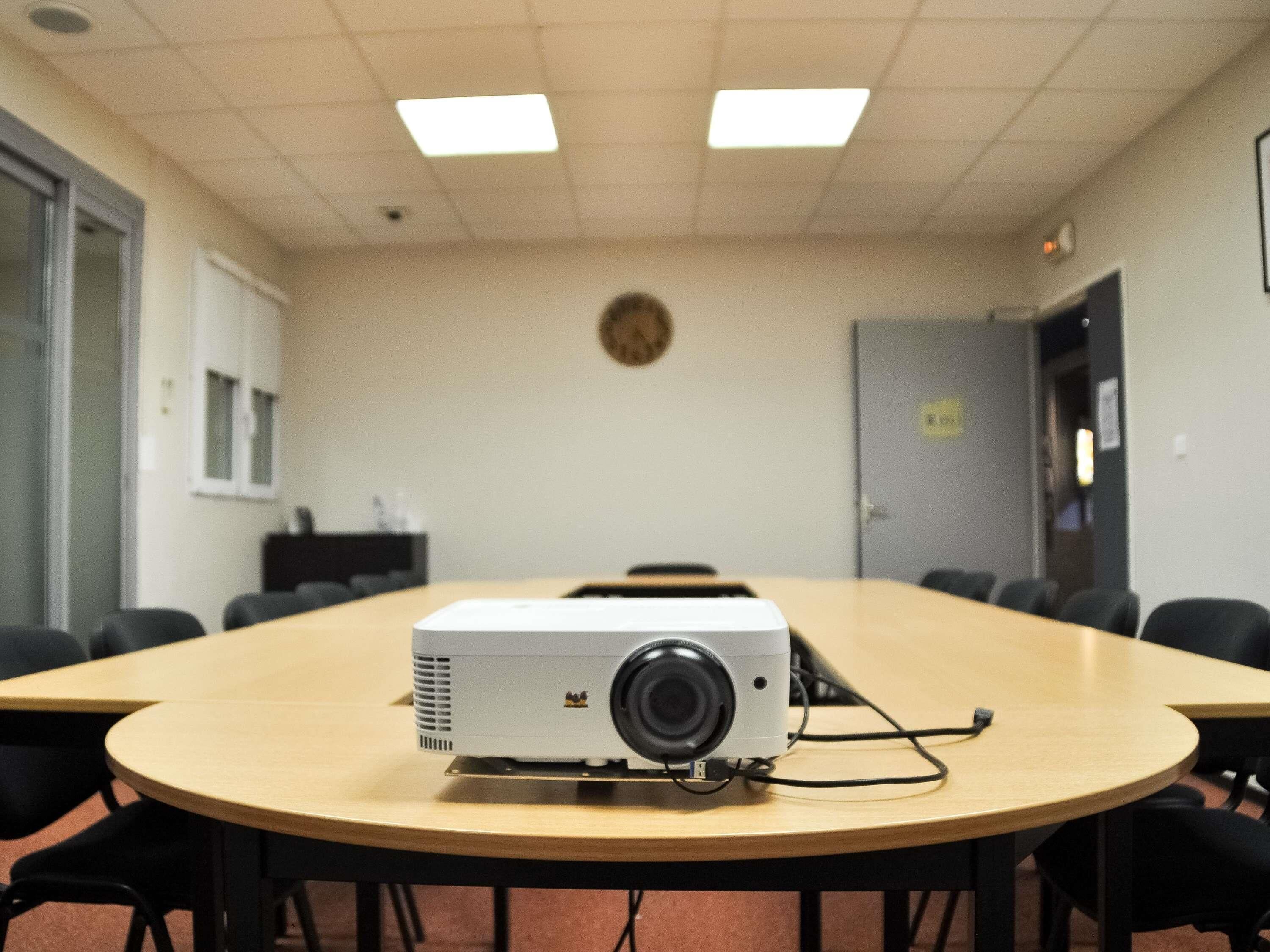 Un salón de conferencias con una gran mesa, sillas, un proyector y un reloj en la pared.