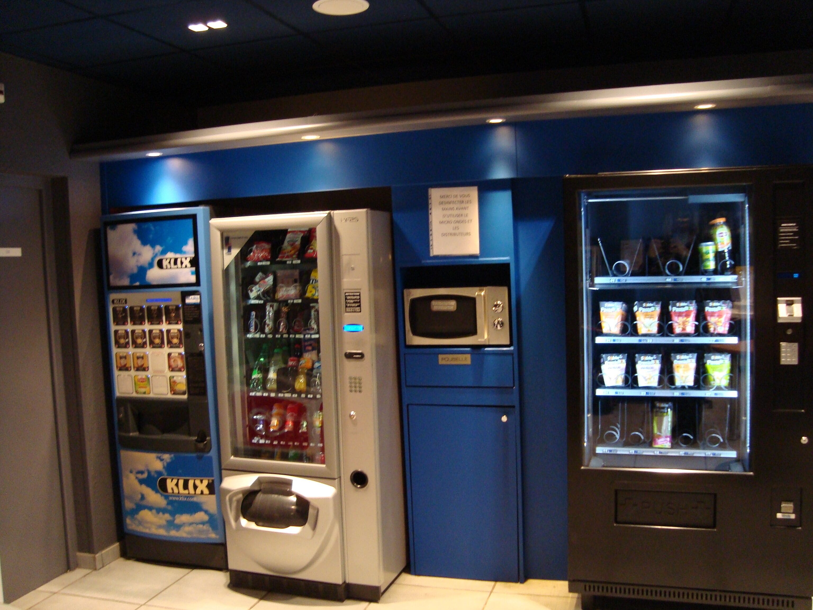 Una hilera de máquinas expendedoras con diversos aperitivos y bebidas, un microondas y un refrigerador pequeño.