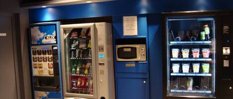 Una hilera de máquinas expendedoras con diversos aperitivos y bebidas, un microondas y un refrigerador pequeño.