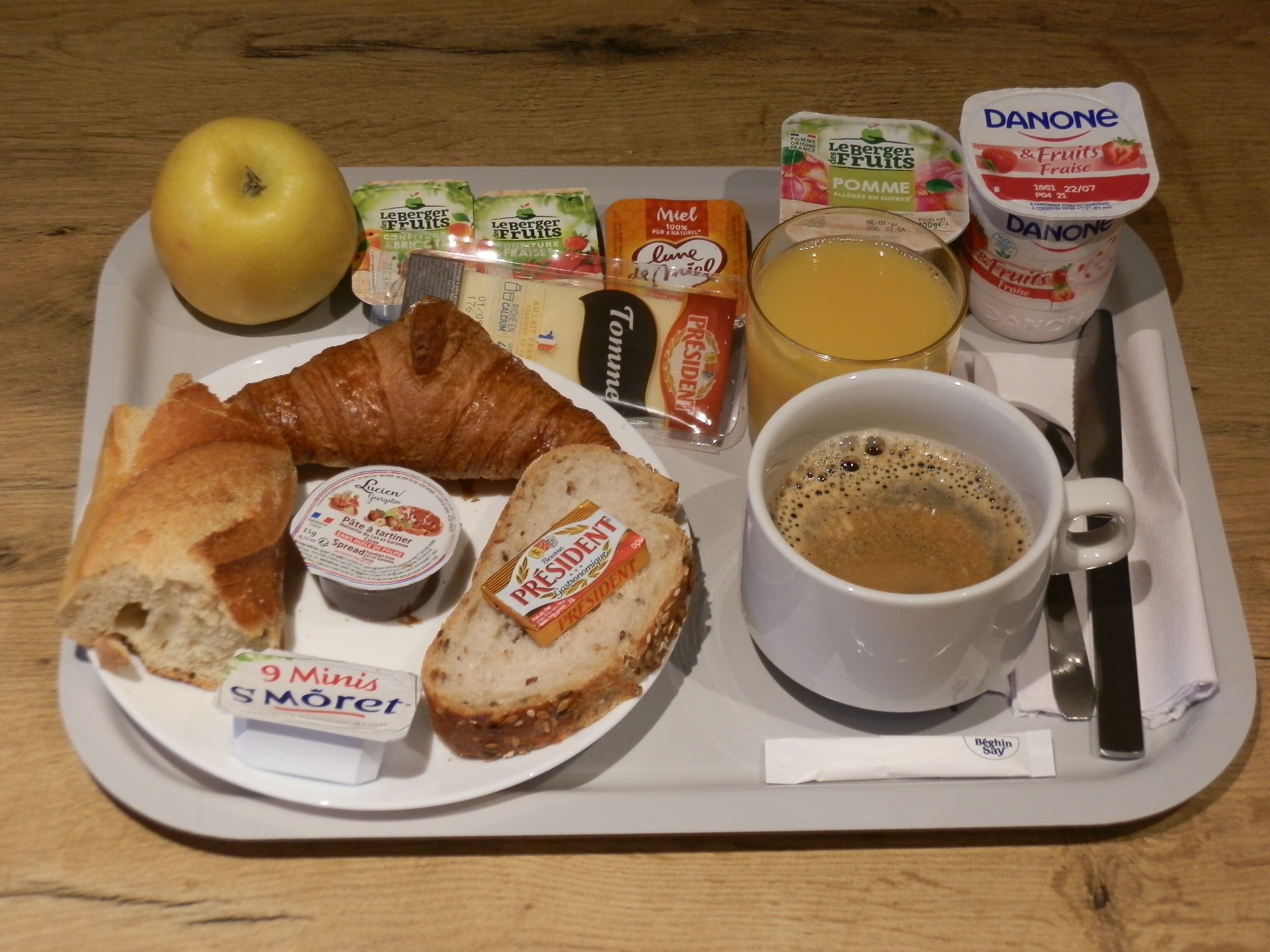 A breakfast tray with croissant, bread, butter, jam, yogurt, juice, and coffee.