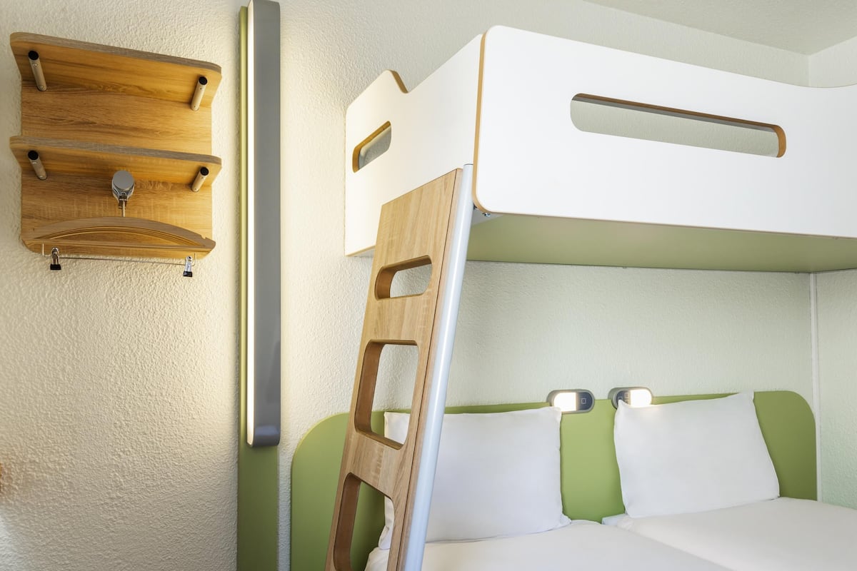 A bunk bed with a ladder, a wooden shelf, and a wall-mounted light fixture.