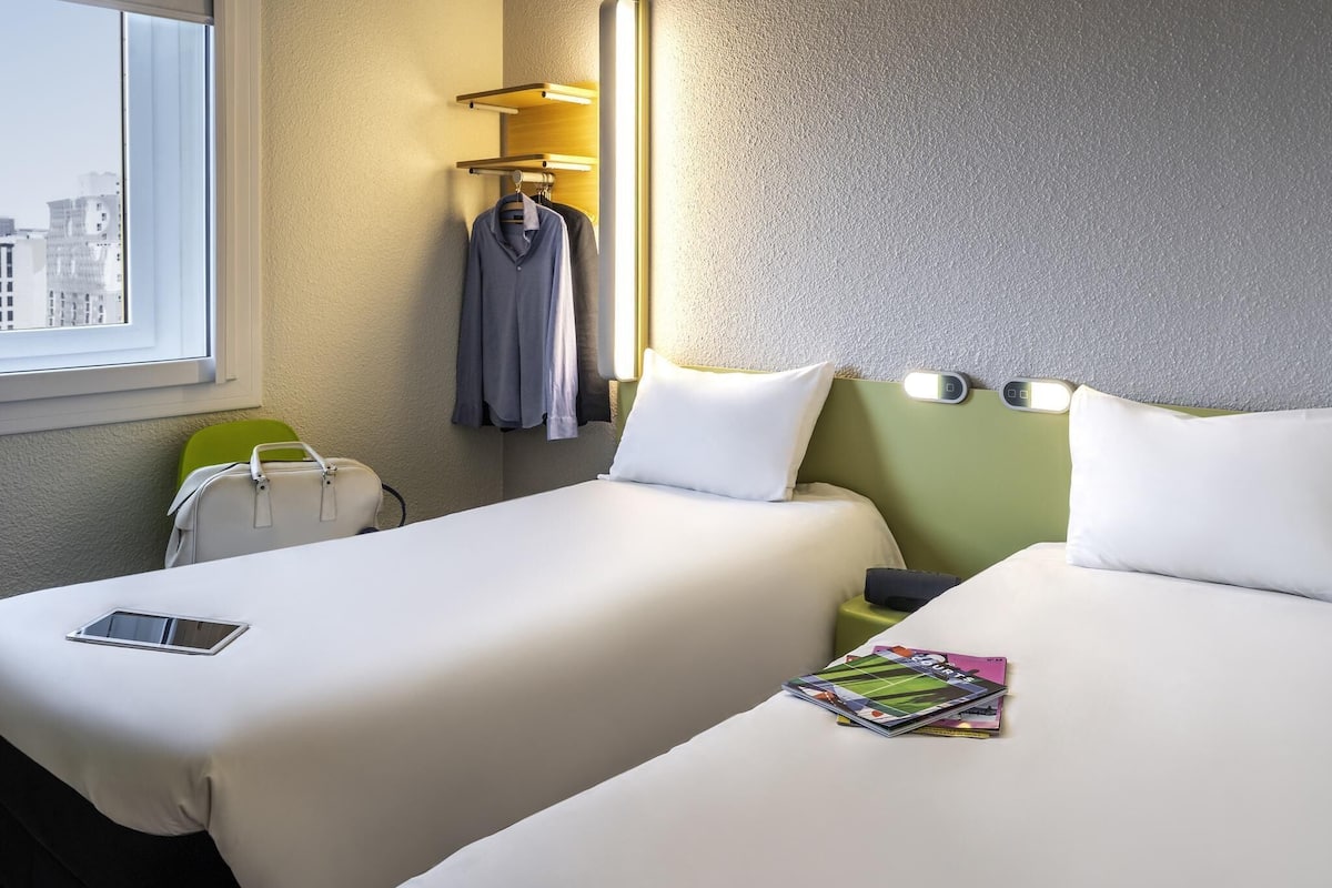 Une chambre d’hôtel avec deux lits, une fenêtre, un porte-manteau mural, une table de chevet avec une lampe, et une vue sur les bâtiments environnants.