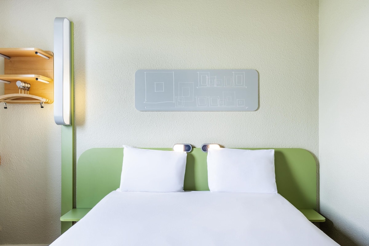 A neatly made bed with white linens, a green headboard, and a wall-mounted light fixture.