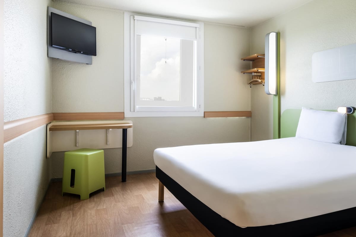 Een hotelkamer met een bed, een aan de muur bevestigde televisie, een raam, een klein krukje en een nachtkastje.