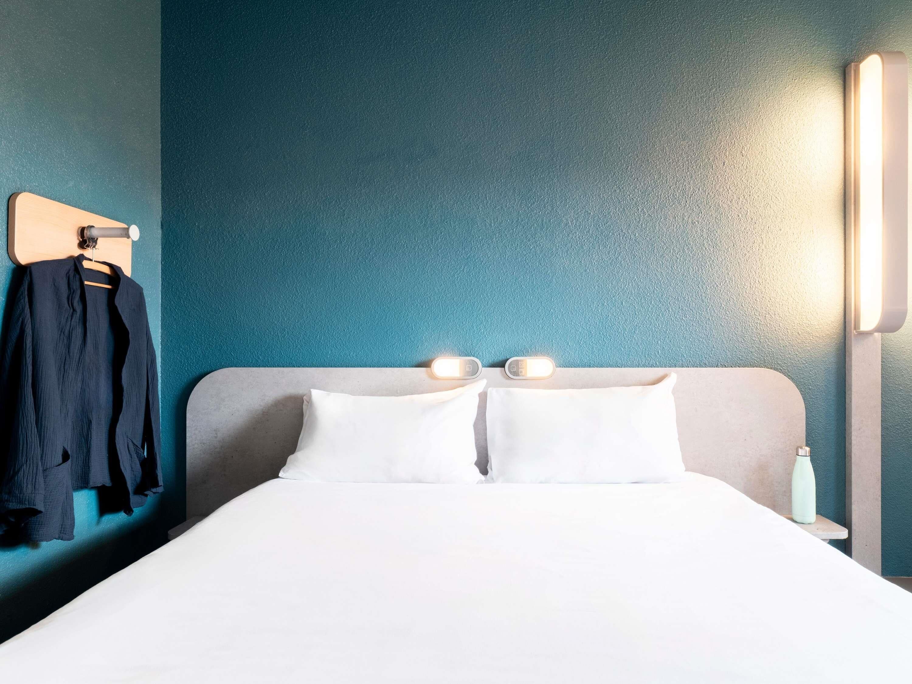 A hotel room with a bed, two bedside lamps, a nightstand, a water bottle, and a coat rack with a jacket.