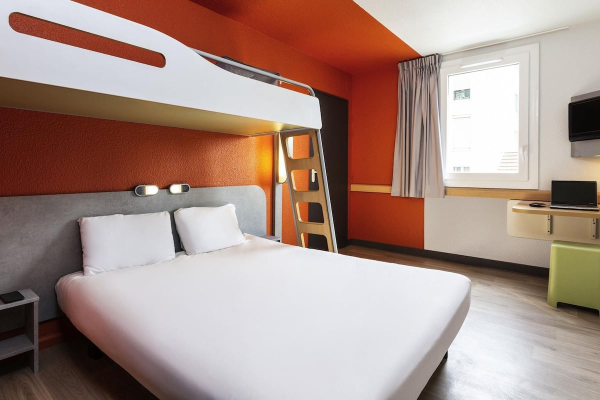 Ein modernes Hotelzimmer mit einem Stockbett, einem Schreibtisch mit einem Laptop, einem Nachttisch und einem Fenster mit Vorhängen.