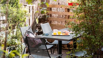 Un área de comedor al aire libre con sillas de metal y una mesa, un cerco de madera y follaje en el fondo.