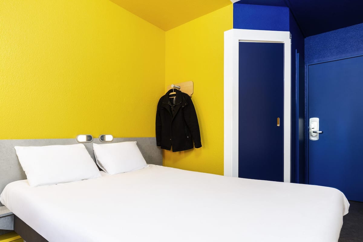 Ein Hotelzimmer mit einem Bett, zwei Nachttischen, einem Kleiderhaken mit einer Jacke und einer blauen Tür.