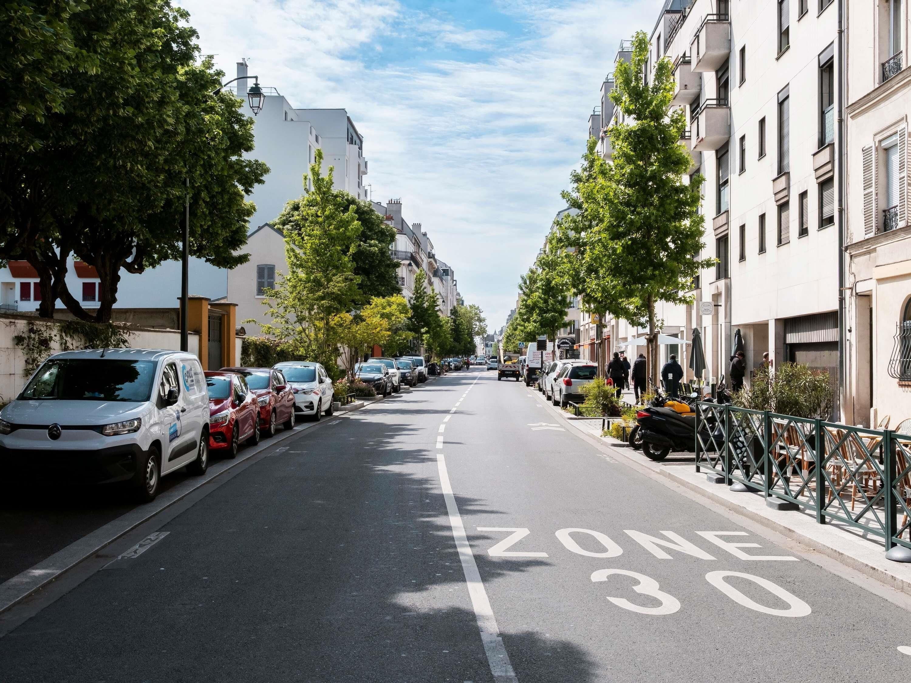 A street lined with parked cars and buildings, a clear sky, and a marked 'ZONE 30' on the road.