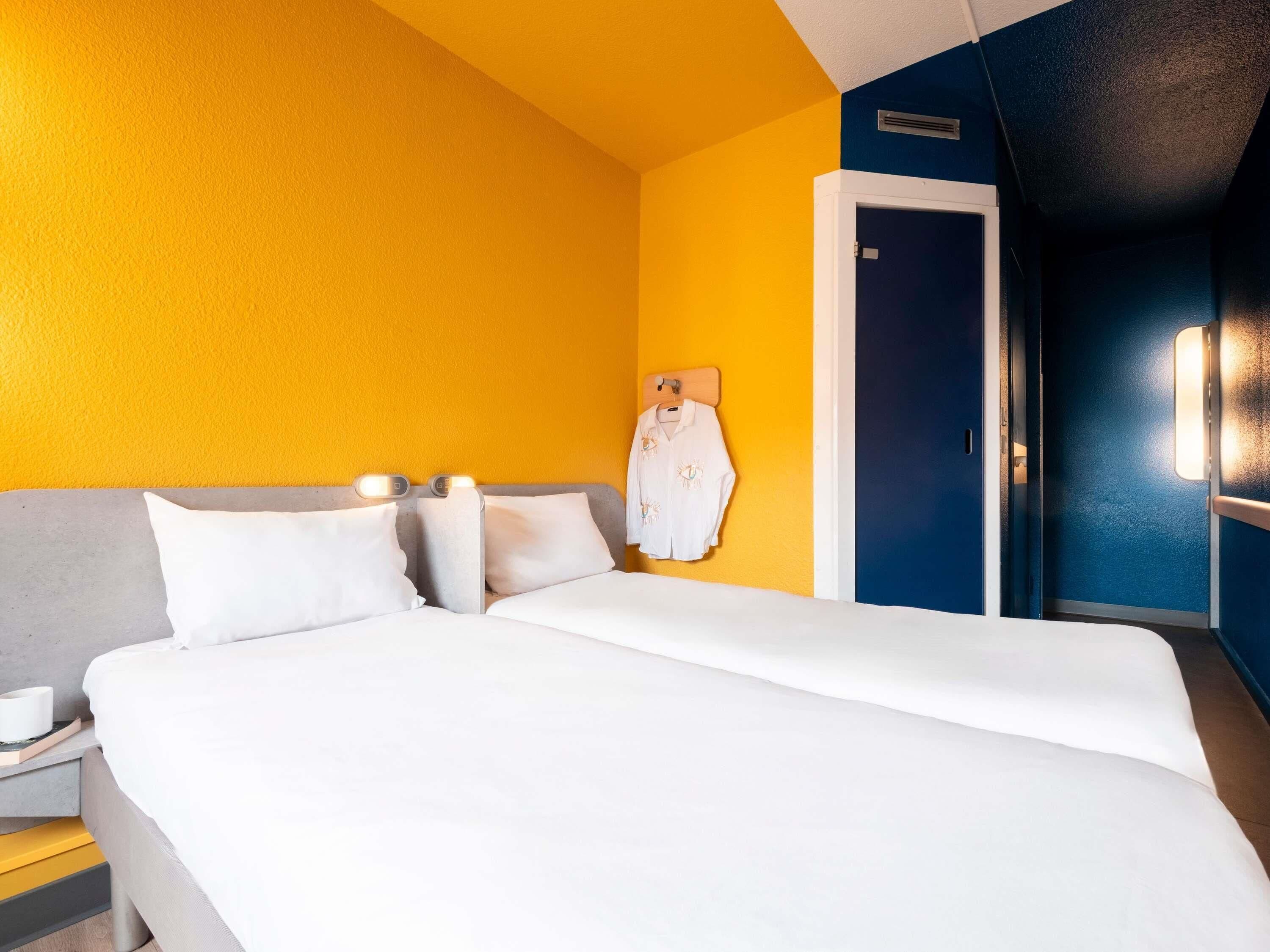 A hotel room with a bed, a nightstand, a door, and a wall with a light switch.