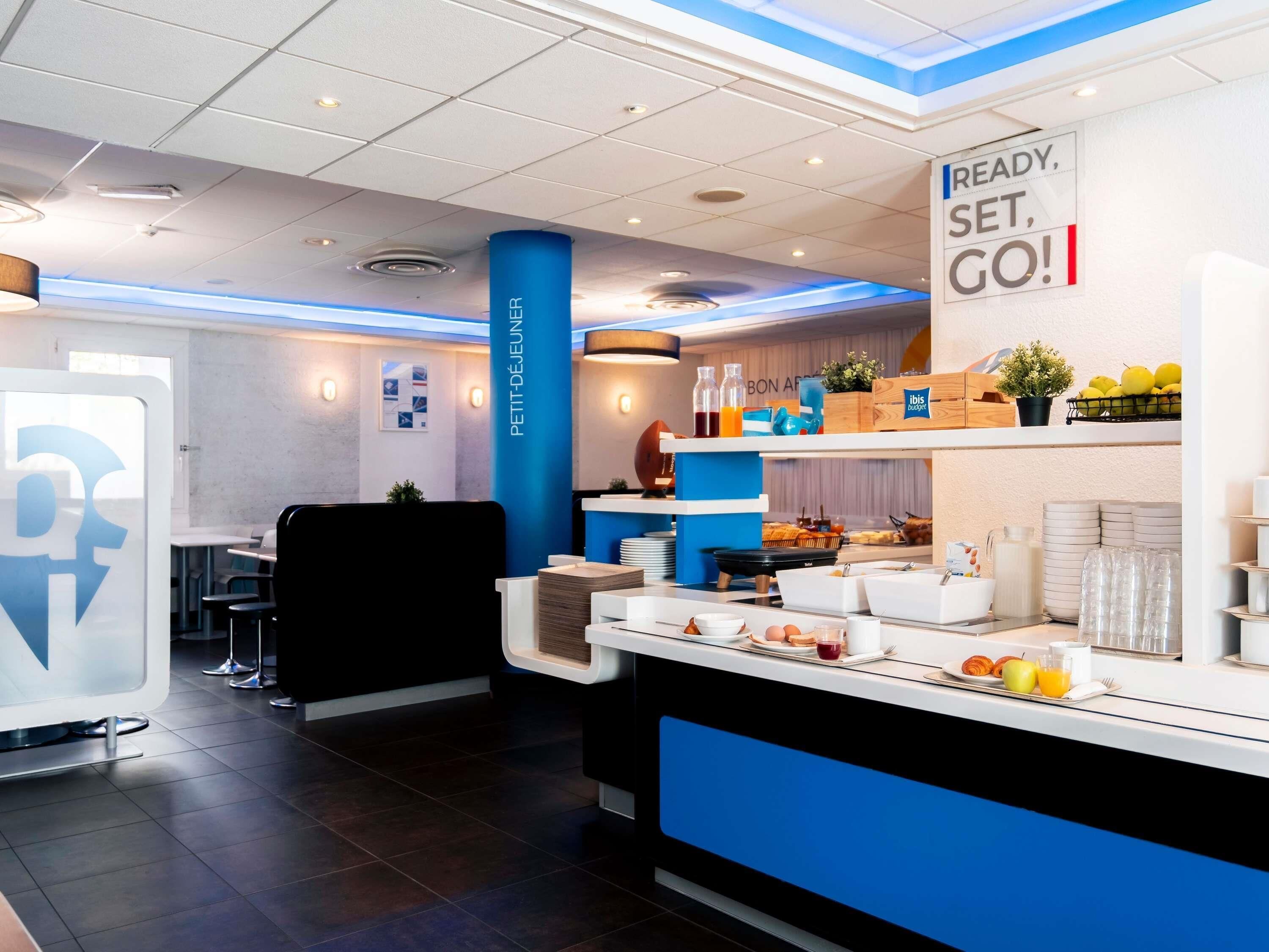 A modern breakfast area with a counter displaying breakfast items, a sign that reads 'READY, SET, GO!', and a blue pillar labeled 'PETIT-DEJEUNER'.