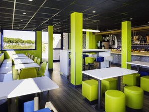 A modern restaurant with high tables and green stools, a bar area, and a view of trees outside. - Ibis Budget Perpignan Nord Rivesaltes (Rivesaltes)