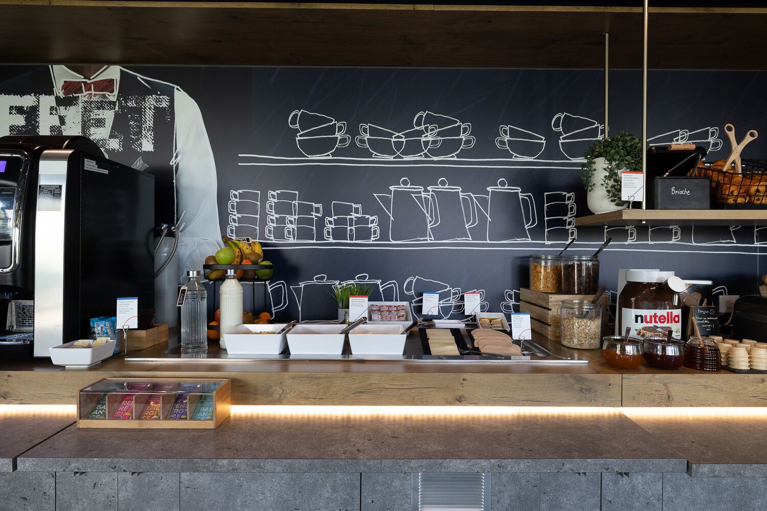 Uma estação de café com um quadro negro exibindo contornos de xícaras de café, uma máquina de café e diversos alimentos, como frutas e acompanhamentos.