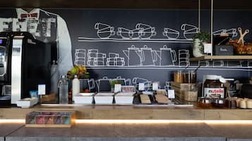 Uma estação de café com um quadro negro exibindo contornos de xícaras de café, uma máquina de café e diversos alimentos, como frutas e acompanhamentos.