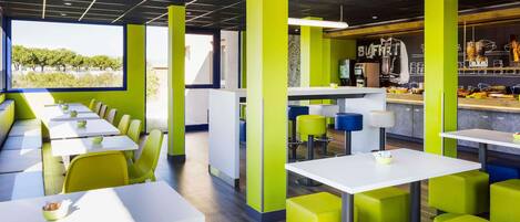 A modern restaurant with high tables and green stools, a bar area, and a view of trees outside.
