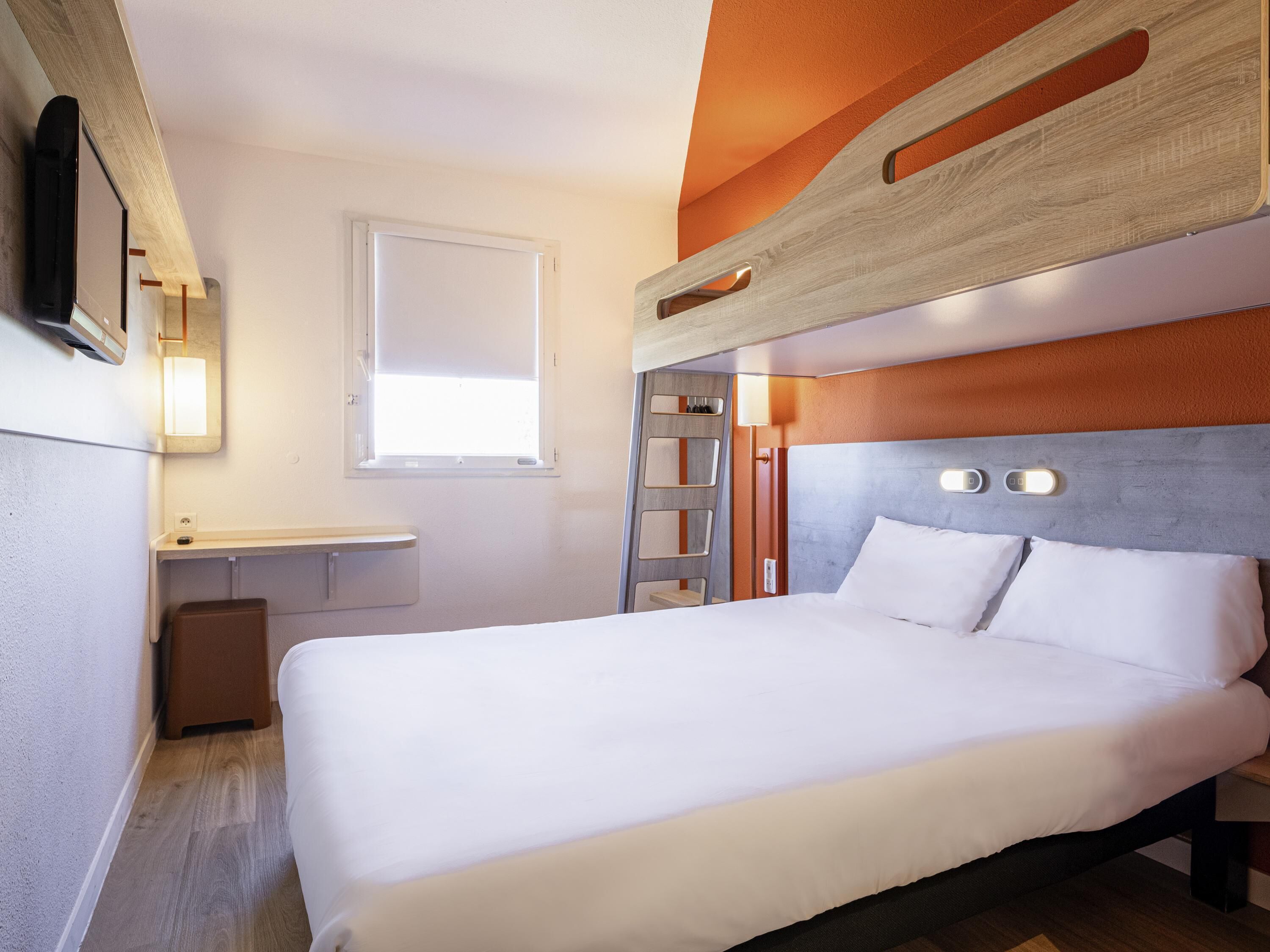 Une chambre d’hôtel avec un lit superposé, une table de chevet, un petit bureau, une chaise, une fenêtre et un téléviseur fixé au mur.