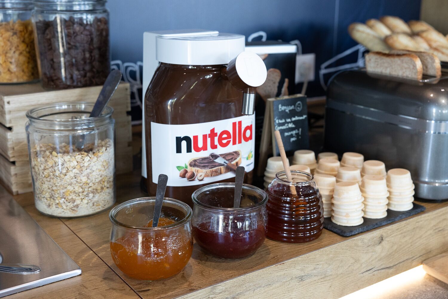 Un frasco de Nutella, frascos de mermelada y miel sobre una encimera de madera.
