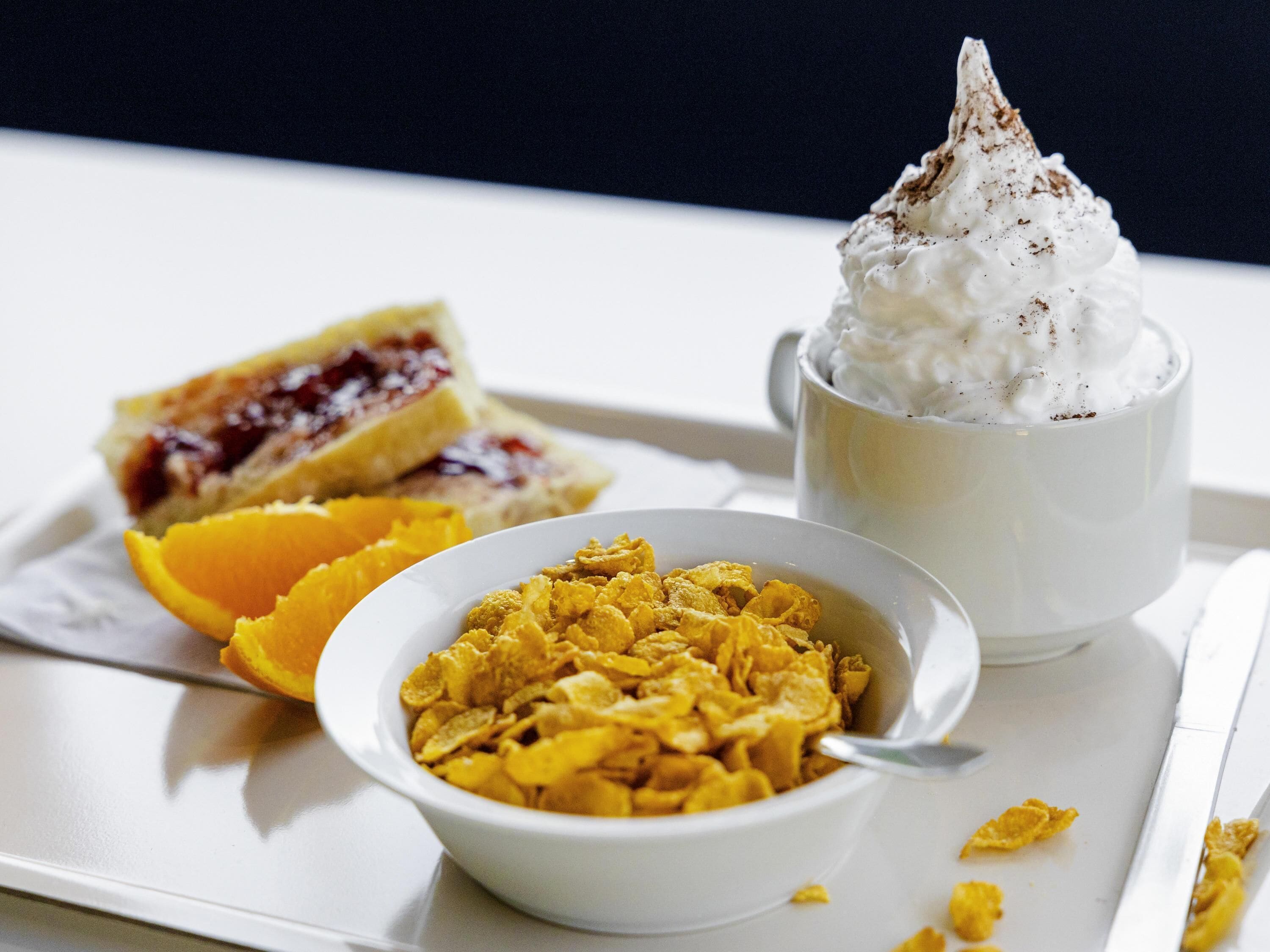 A tray with a cup of whipped cream, a bowl of cereal, a slice of toast with jam, and an orange half.