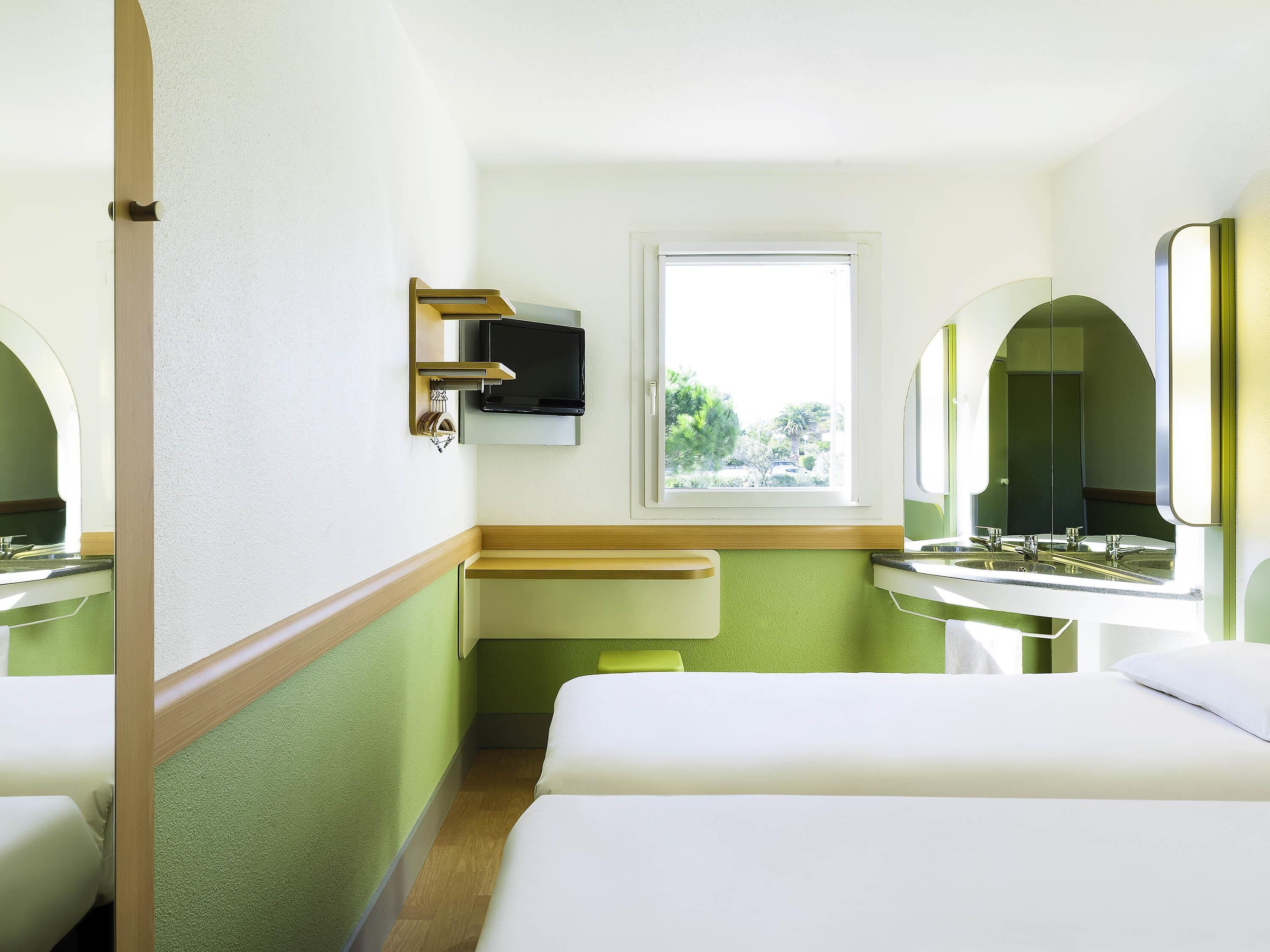 Une chambre d’hôtel avec deux lits, une télévision, une fenêtre et une salle de bain avec un miroir.