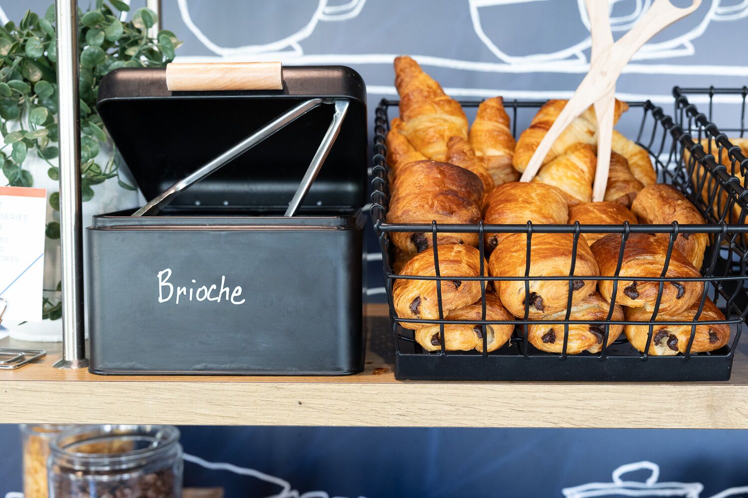 A black metal box labeled "Brioche" with a wooden handle, containing a basket of golden-brown brioche pastries.