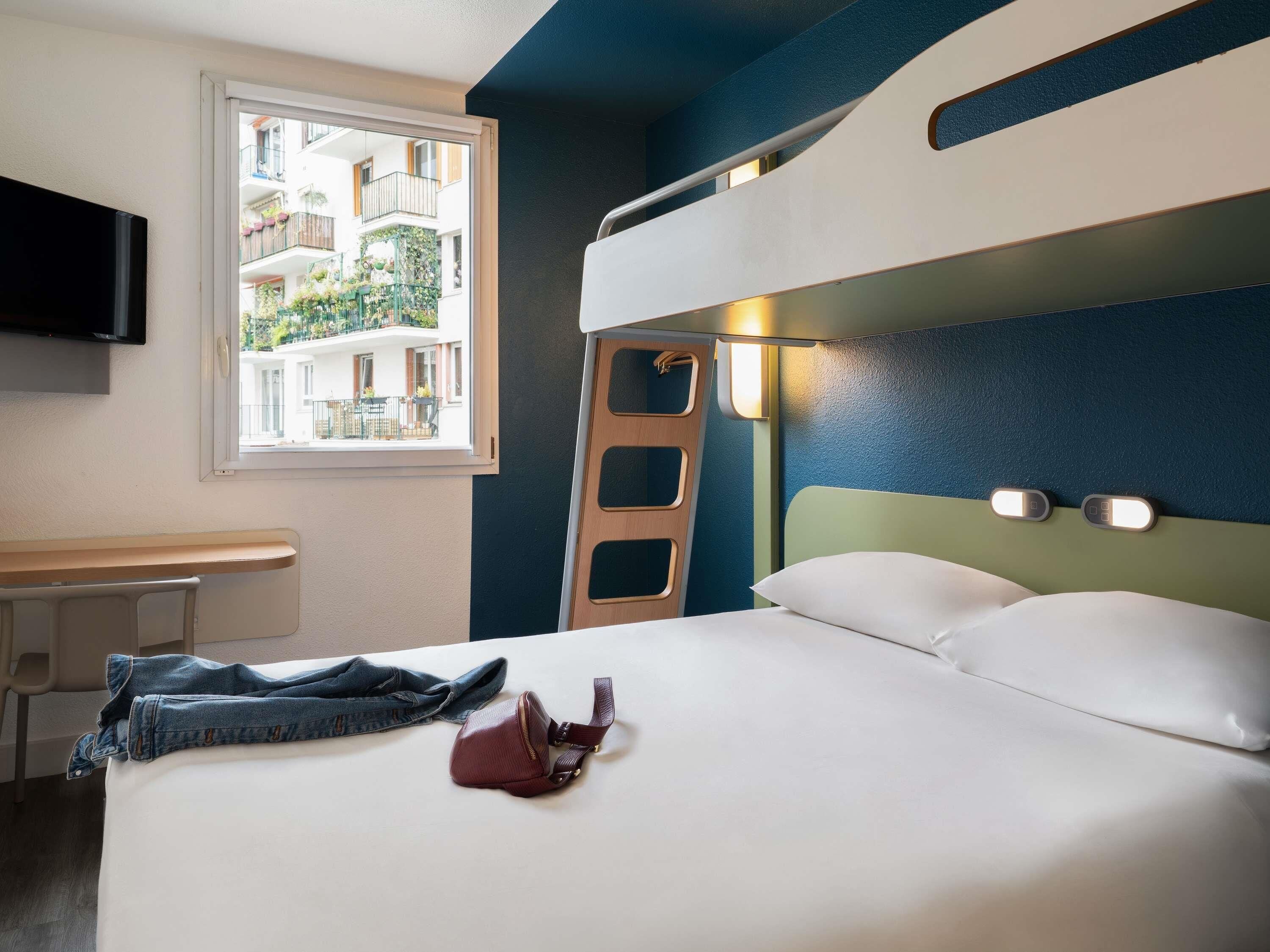 A hotel room with a bunk bed, a desk, a chair, a window with a view of buildings, and a TV mounted on the wall.