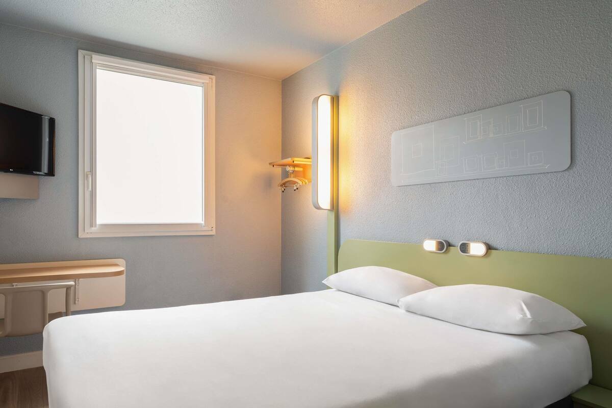 A hotel room with a bed, a bedside table, a TV, a window, and a wall-mounted light fixture.