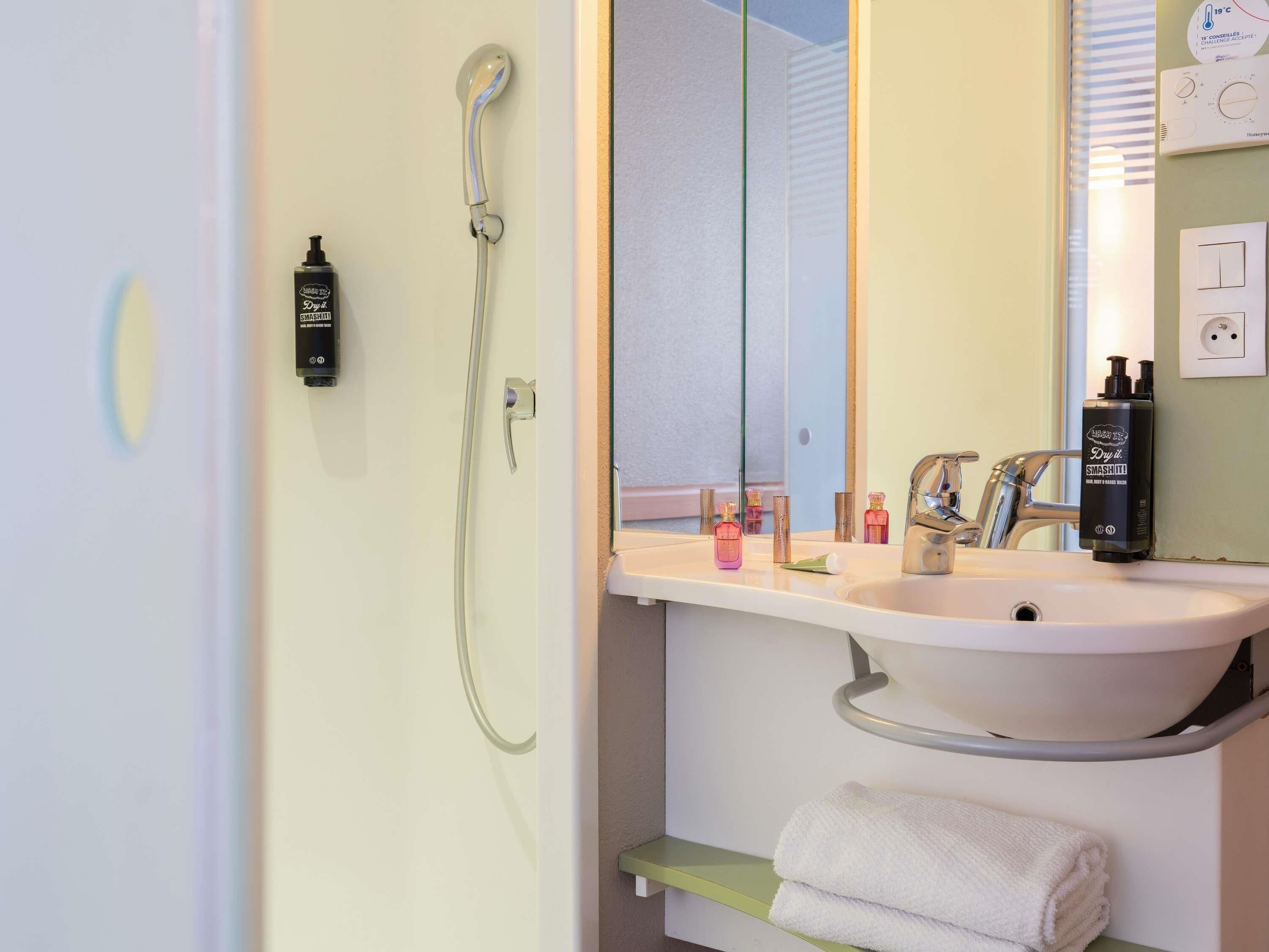 Un baño con lavamanos blanco, cabezal de ducha de mano, toallero y espejo.