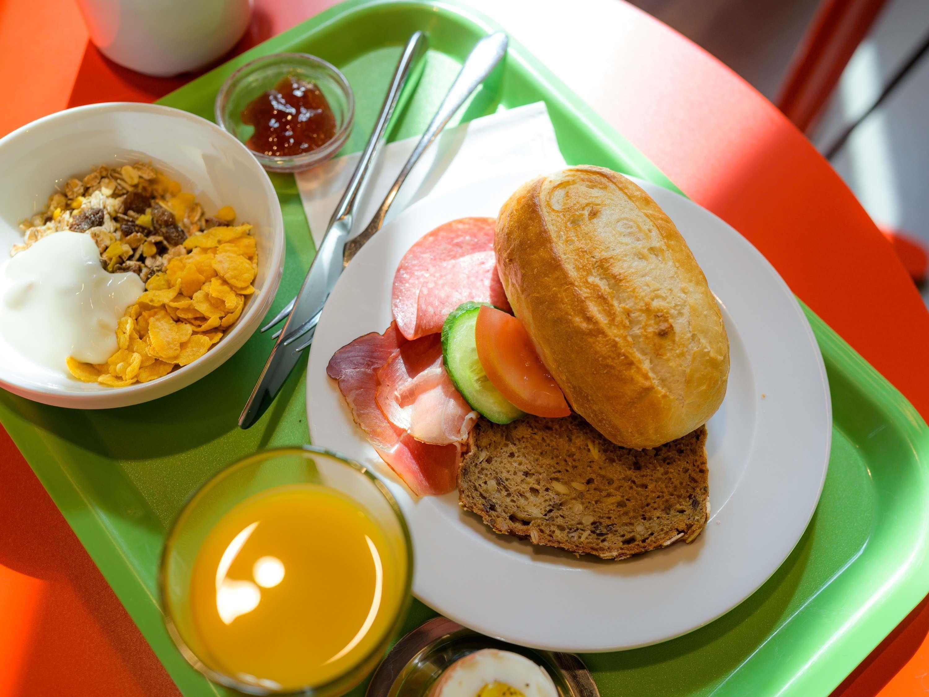 A breakfast tray with cereal, a sandwich, a slice of bread, a small bowl of jam, and a glass of orange juice.