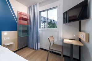 Une chambre d’hôtel comprenant un lit, un bureau, une chaise, une télévision, une fenêtre donnant sur des immeubles et un rideau.
