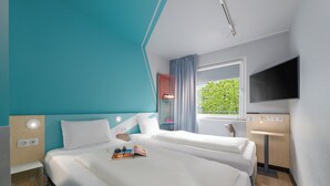 A hotel room with two beds, a TV, a window with curtains, and a small desk area. - ibis budget Leipzig City (Leipzig)