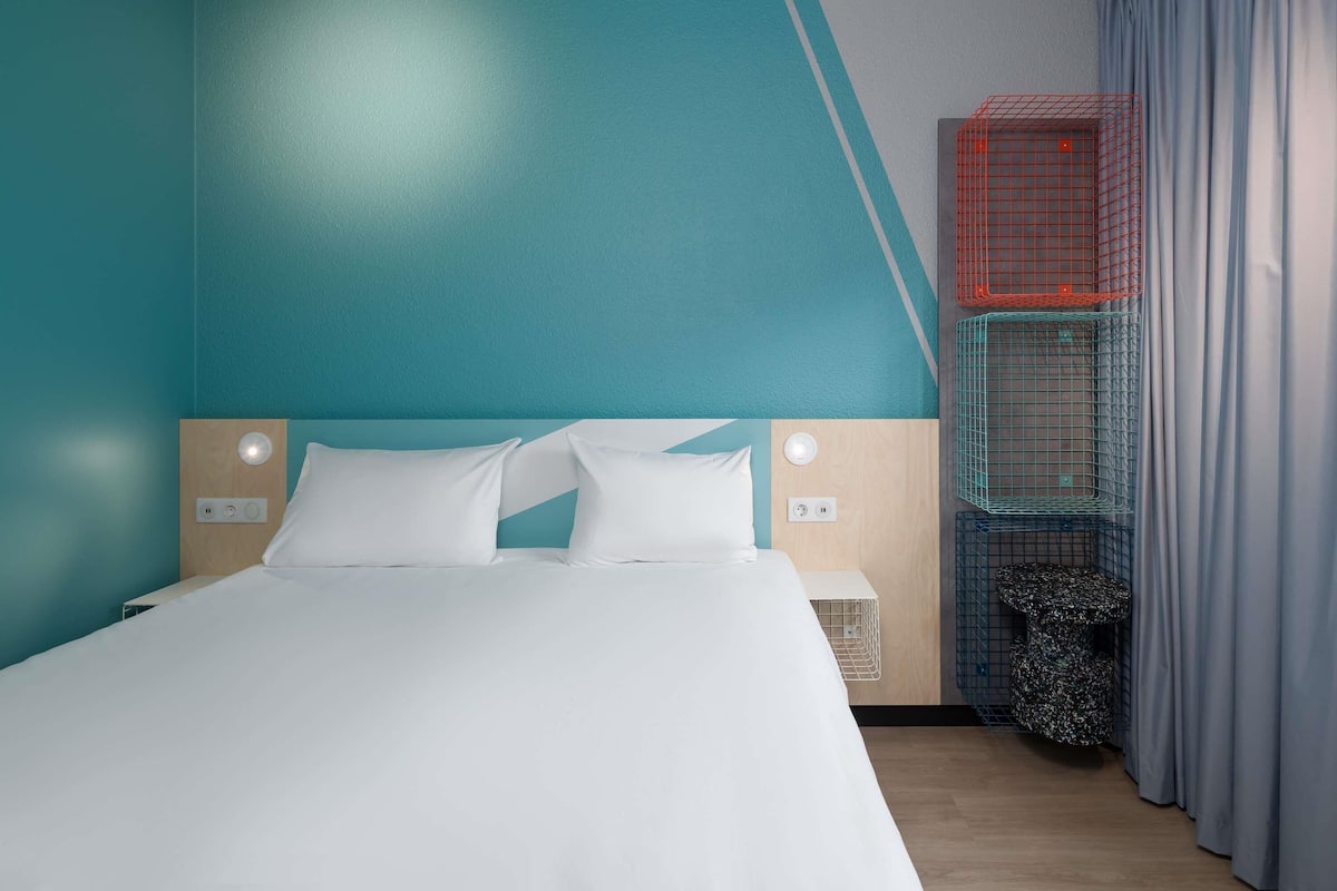A modern hotel room with a bed, white bedding, a headboard with a light fixture, a small bedside table, and a wall-mounted shelf.