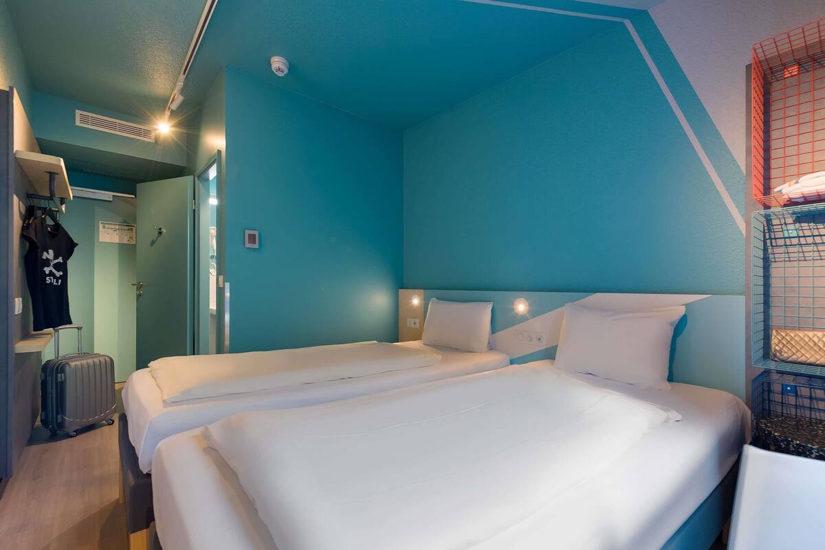 A hotel room with two beds, a wardrobe, a suitcase, and a bunk bed with a storage box.