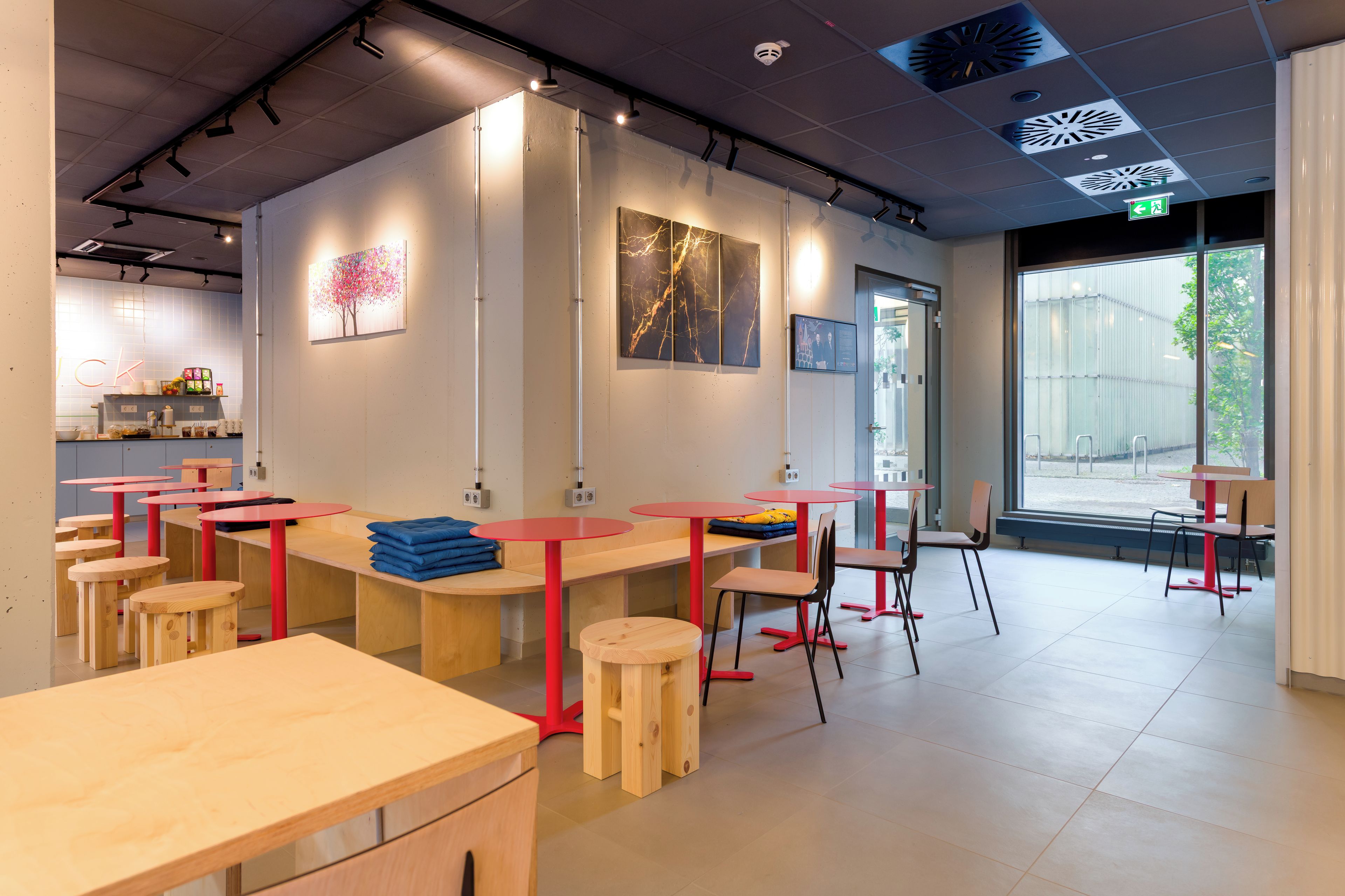 Interior de una cafetería moderna con mesas de madera y sillas rojas, obras de arte en las paredes y un amplio ventanal con vistas al exterior.